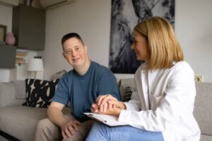 Happy man with special needs having a support by his home caregiver in the living room and looking at camera.